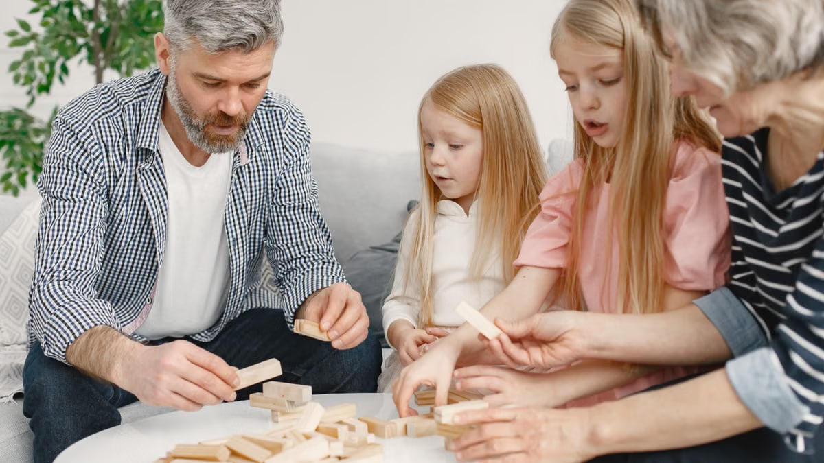 Four family members of different ages engage with toy blocks on a table indoors.